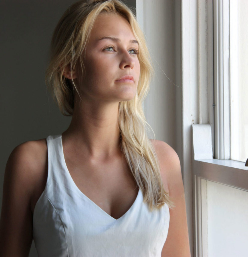 Woman in white tank top looking contemplatively out a window.
