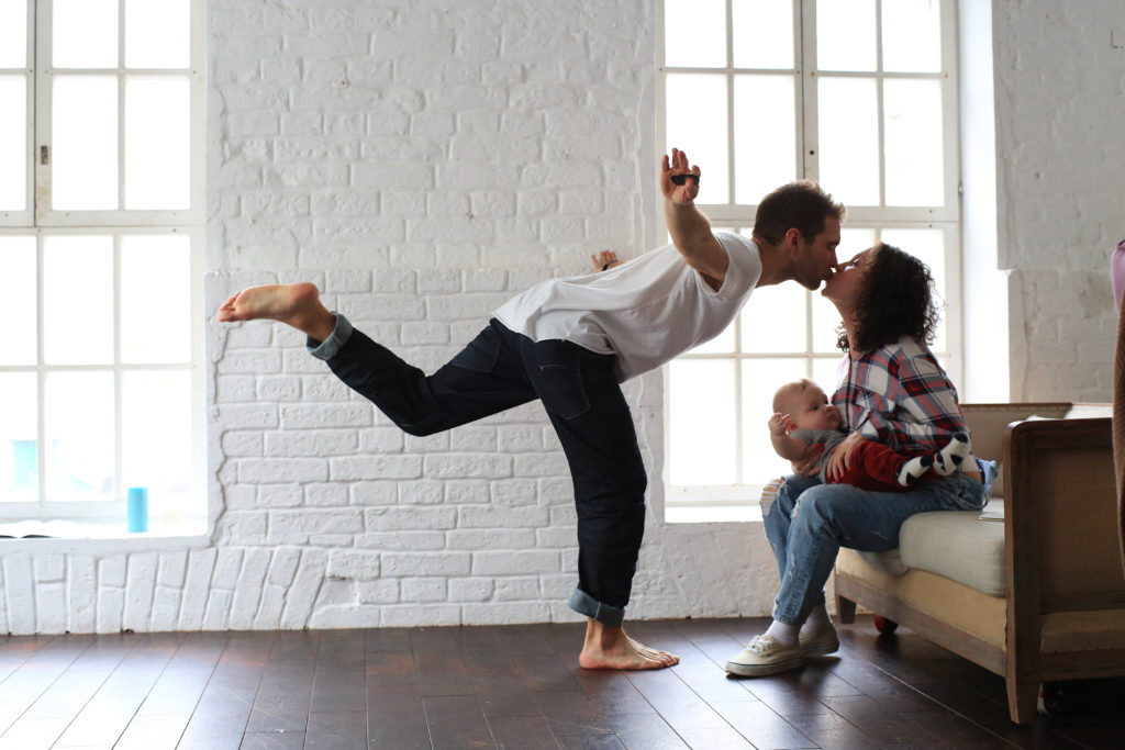 Homem se equilibrando em um pé enquanto tira uma selfie e beija uma mulher segurando um bebê.