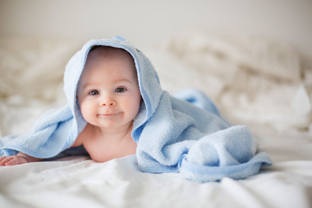 Bambino avvolto in un asciugamano con cappuccio blu sdraiato su un lenzuolo bianco.
