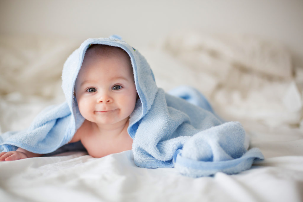 Baby, eingewickelt in ein blaues Kapuzenhandtuch, liegt auf einem weißen Bettlaken.