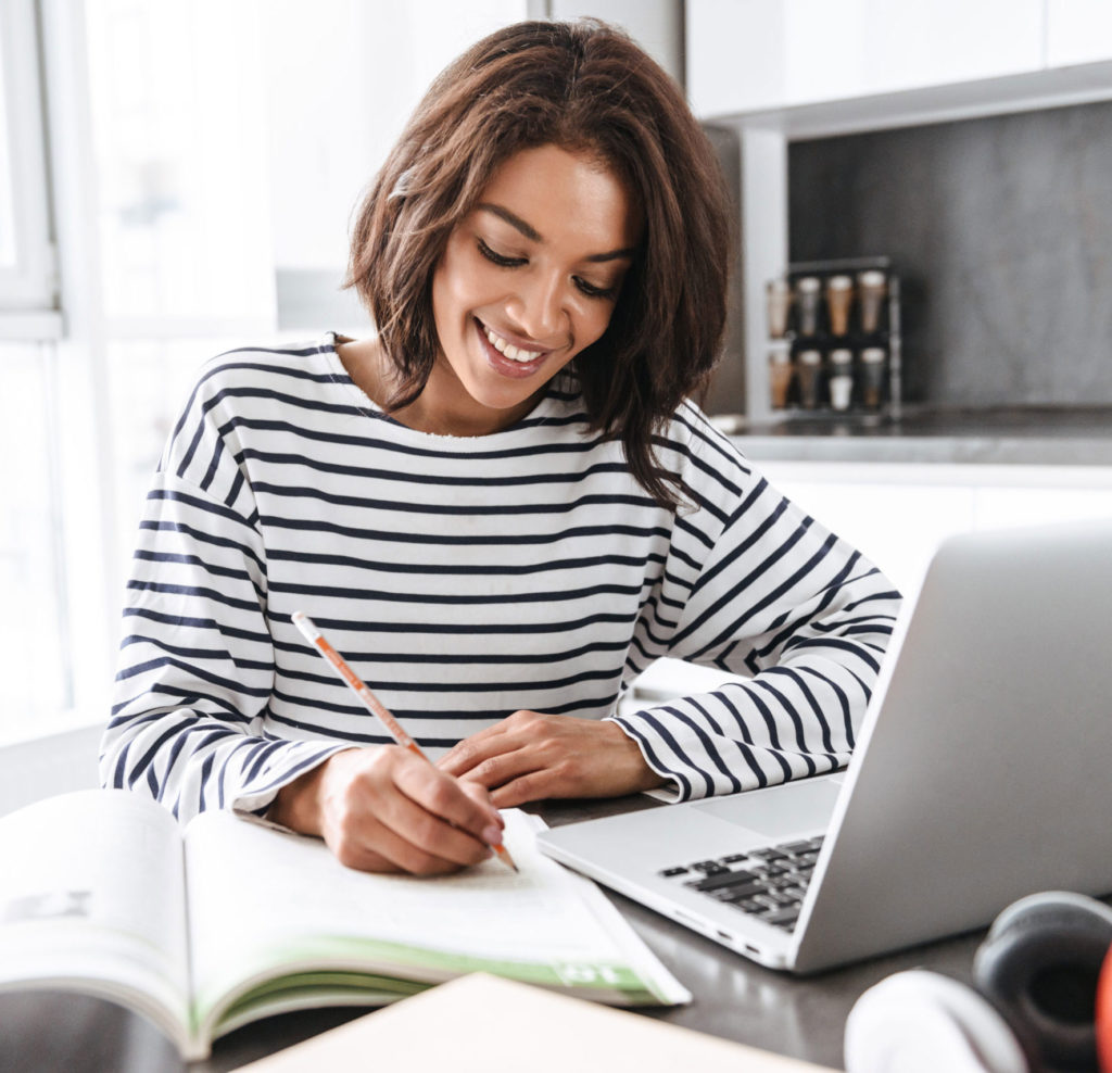امرأة ترتدي قميصًا مقلمًا وتكتب في دفتر ملاحظات وأمامها كمبيوتر محمول مفتوح.