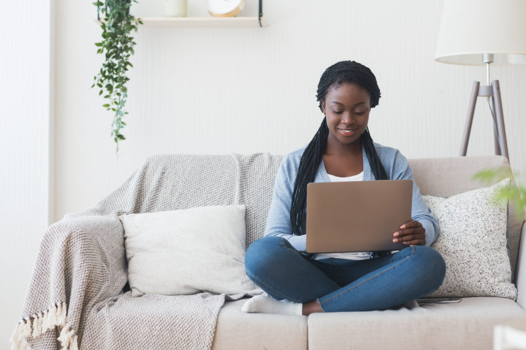 Black millennial woman working with laptop computer on sofa at home