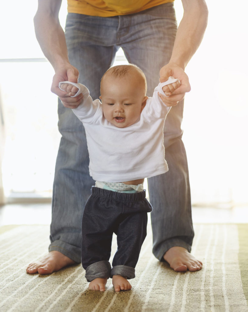 Kleinkind unternimmt unterstützte Schritte, während es sich an den Händen eines Erwachsenen festhält.