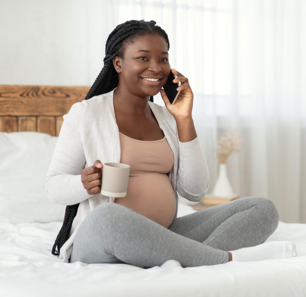 Surrogate mother smiling, talking on the phone and holding a mug, sitting on a bed.