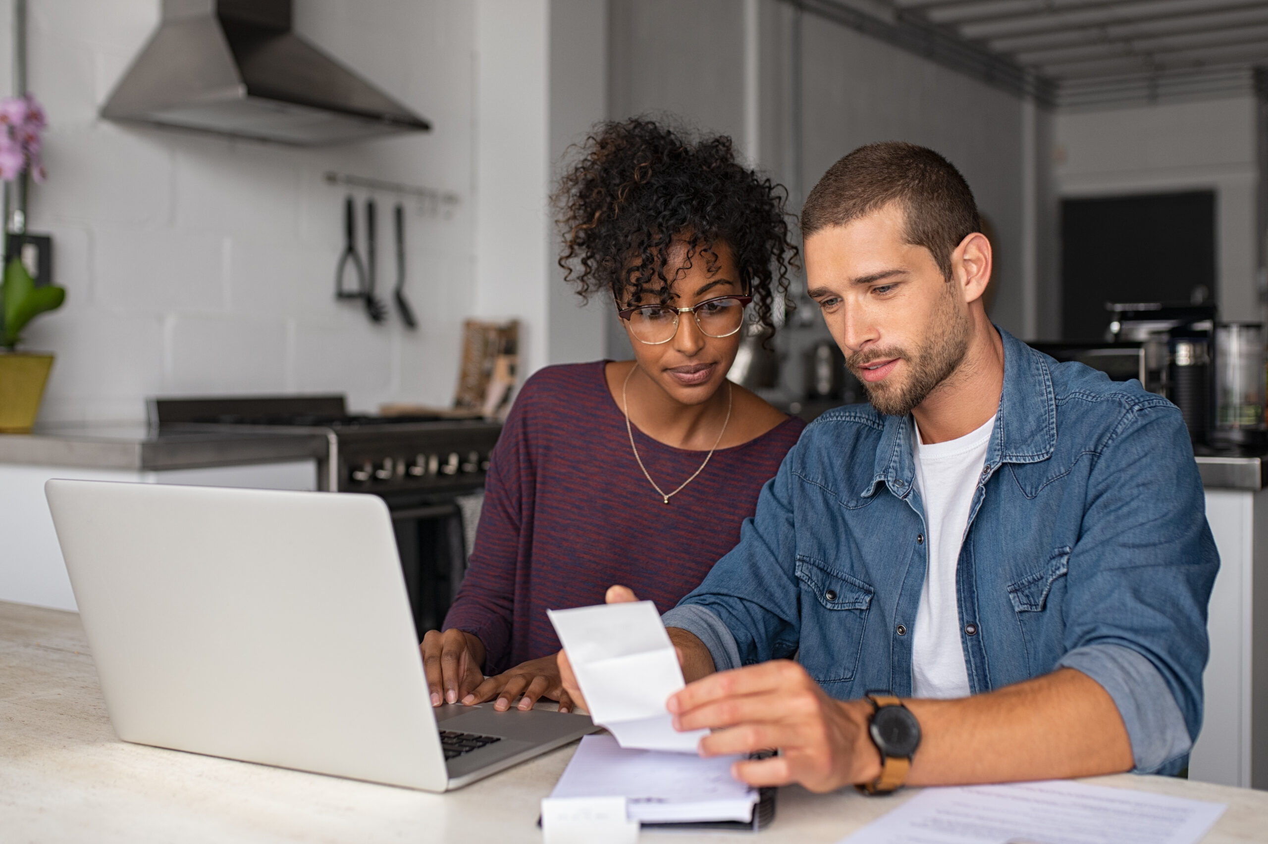 Pareja multiétnica revisando los costos de la subrogación y usando una computadora portátil en la cocina de una casa.