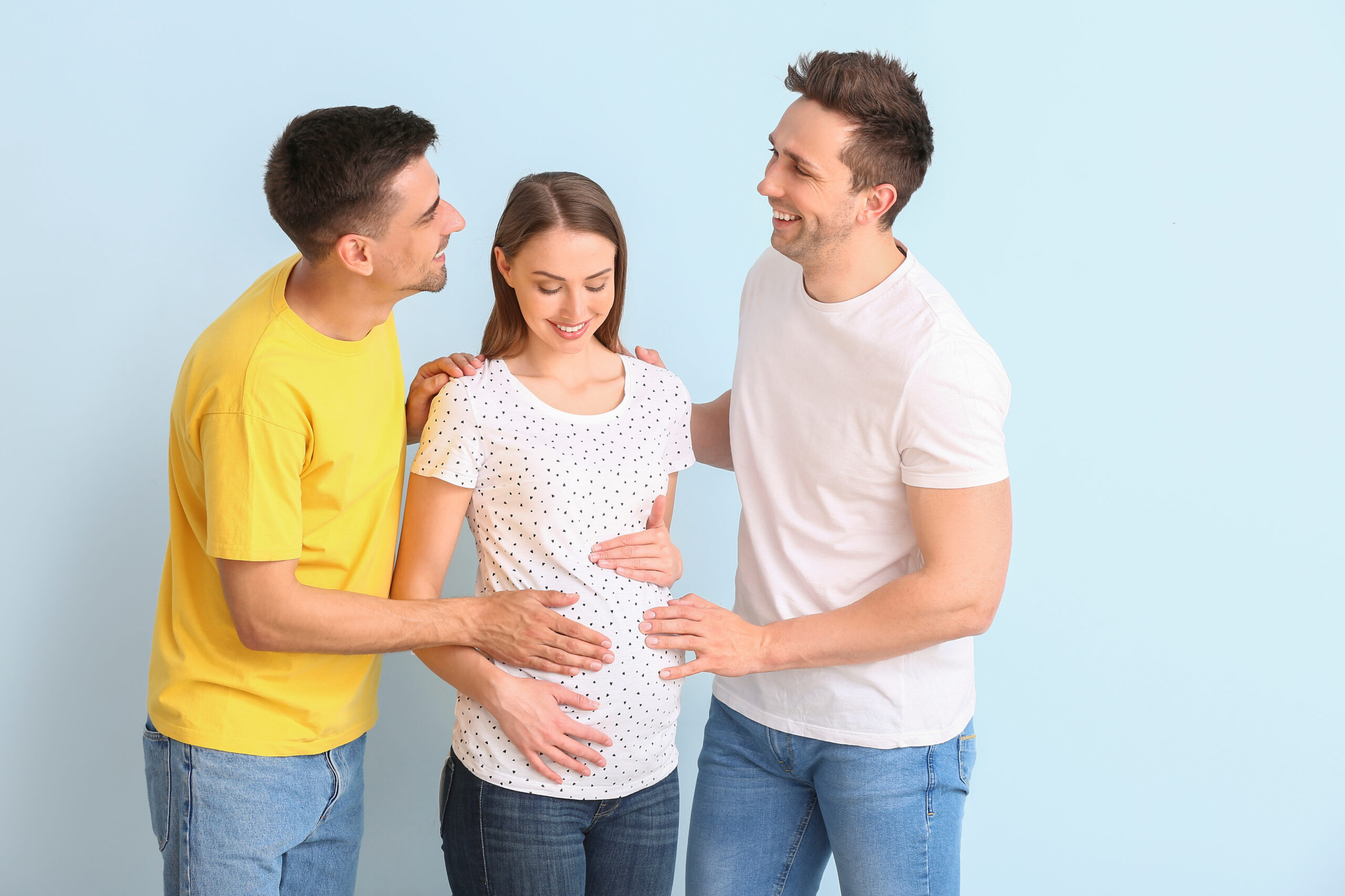 Dois homens e uma mulher grávida sorrindo juntos com as mãos na barriga, sugerindo um acordo de barriga de aluguel para pais LGBTQ.