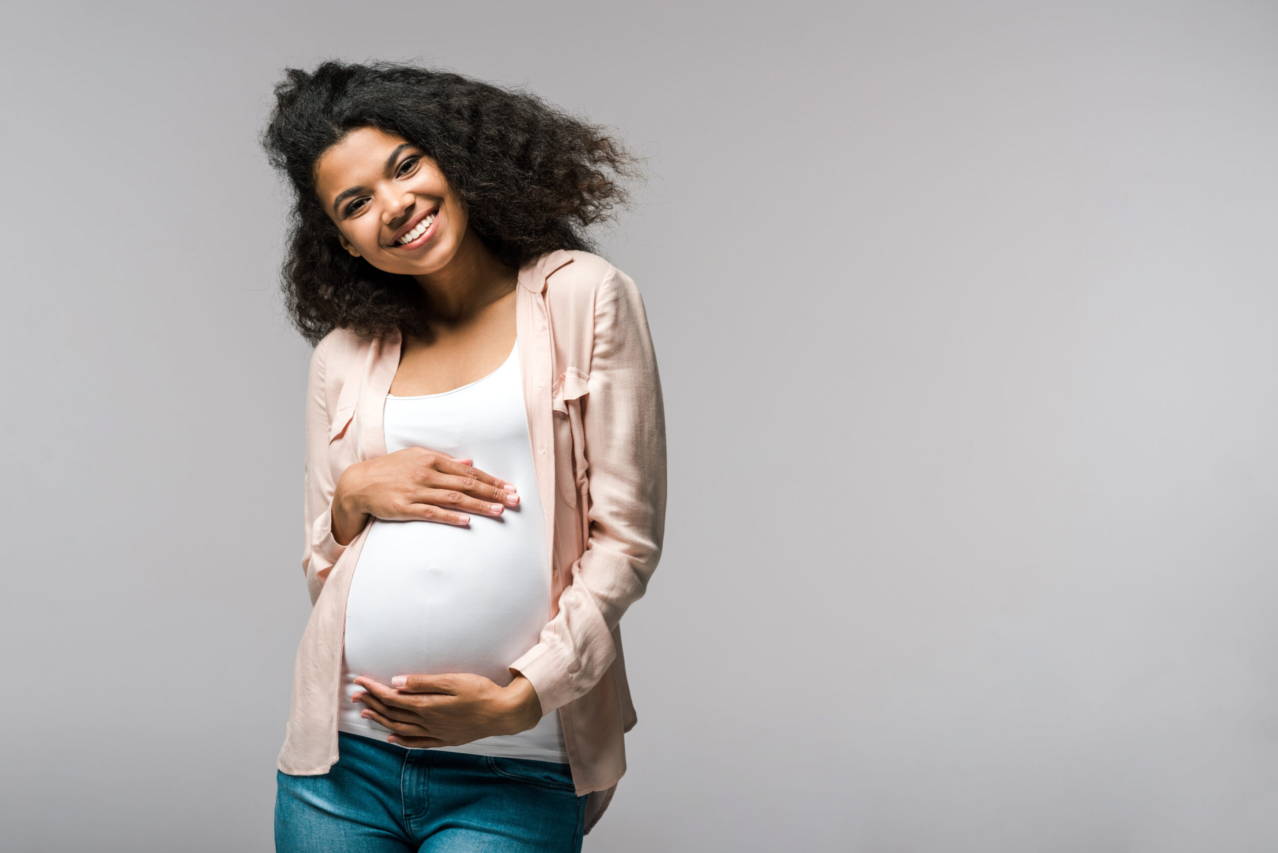 Lächelnde schwangere Frau, die ihren Bauch hält, ein weißes Oberteil und eine leichte Strickjacke trägt.